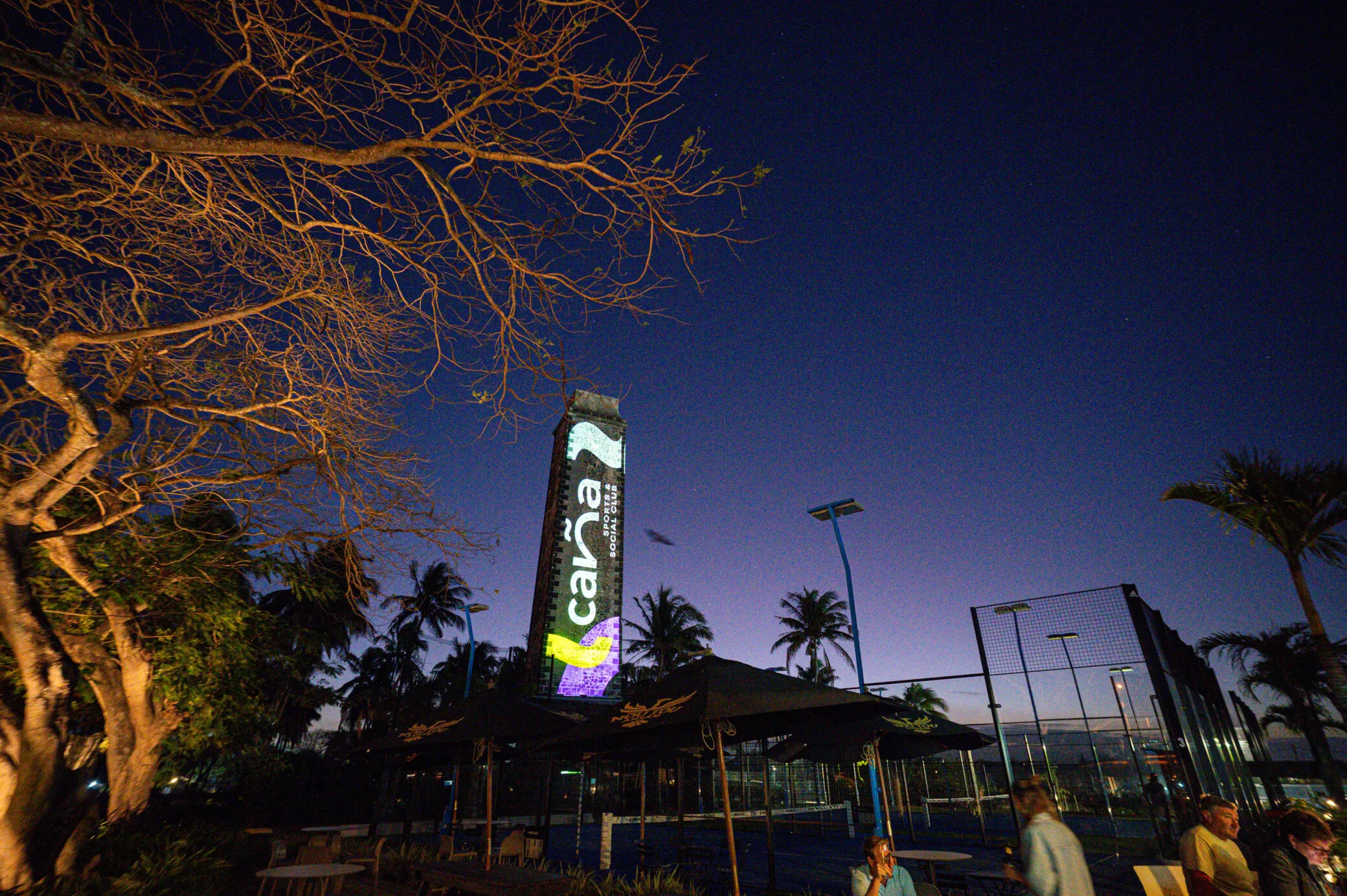 Old chimney tower lit at dusk with the Caña padel and social club logo, beside padel courts, palm trees and a terrace